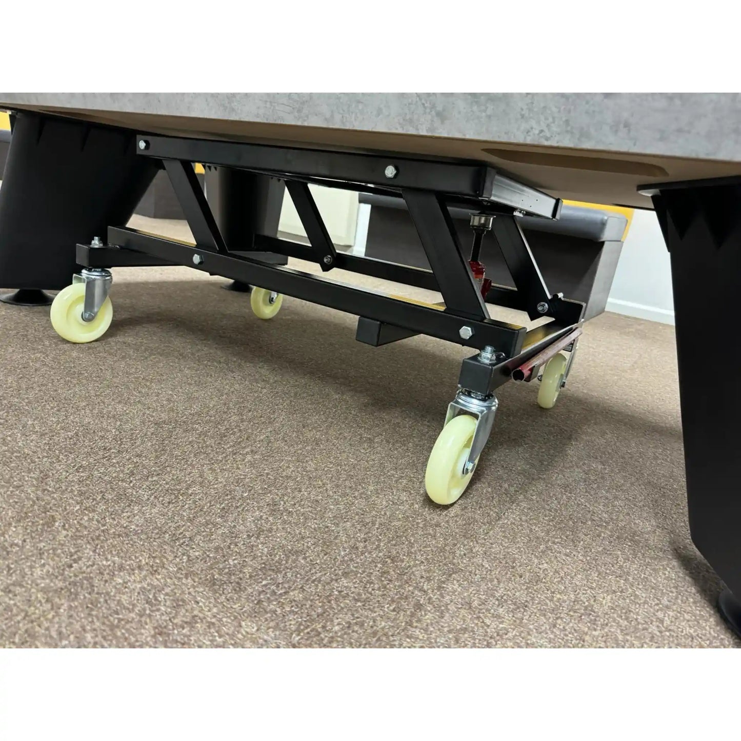 A close-up view of a hydraulic pool table trolley with a black metal support structure and caster wheels, shown on a carpeted floor.