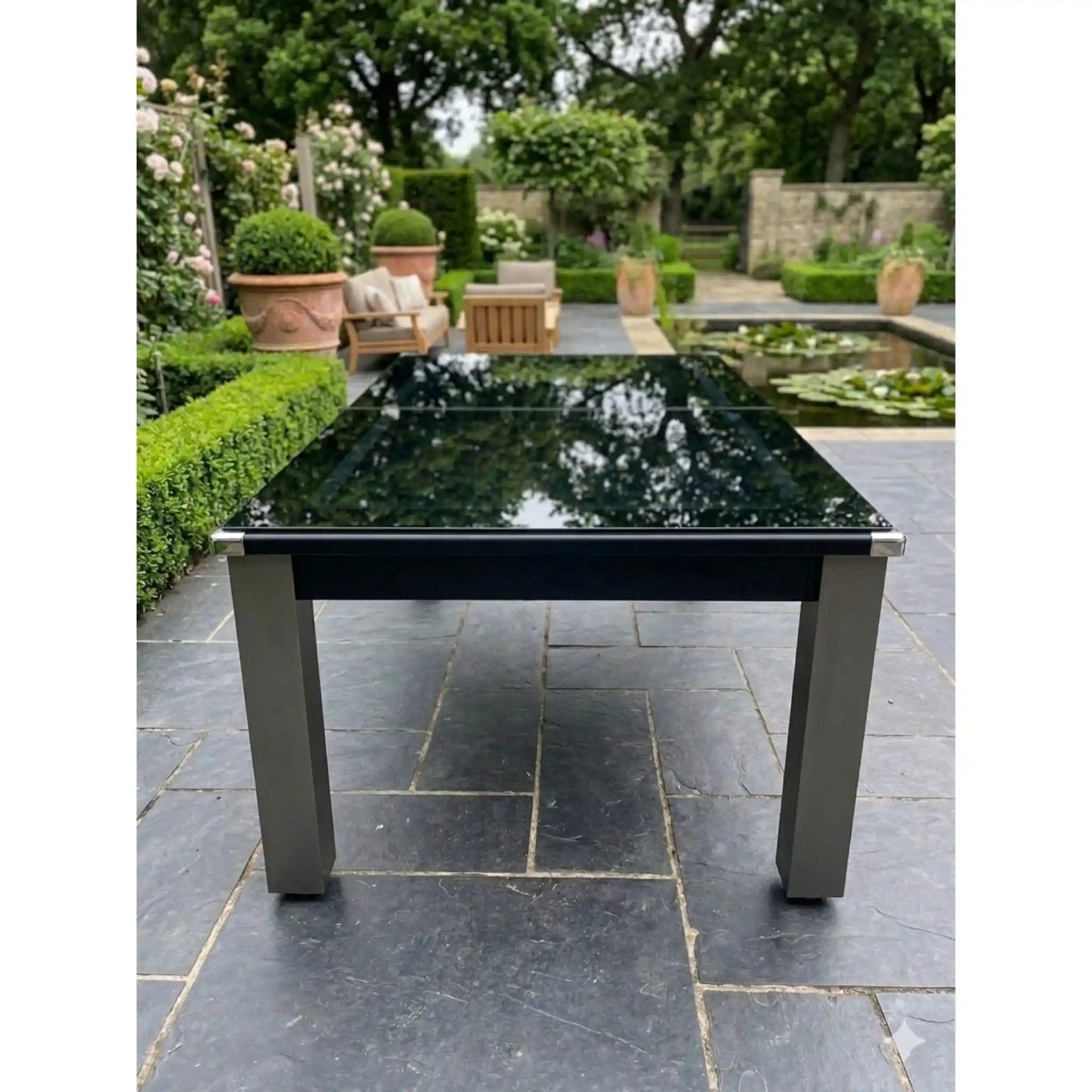 A modern outdoor pool dining table with a glossy black rectangular glass top and a brushed silver metal frame and legs, set on a dark stone patio in a landscaped garden.