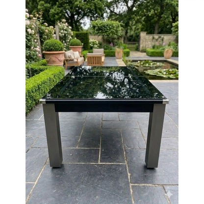 A modern outdoor pool dining table with a glossy black rectangular glass top and a brushed silver metal frame and legs, set on a dark stone patio in a landscaped garden.