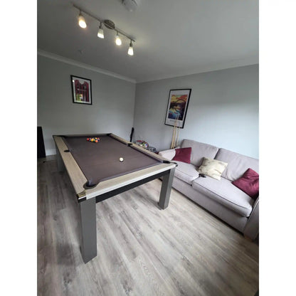 A pool table with a dark brown felt surface and light wood-grain rails, positioned in a home game room next to a light gray sofa with pillows.