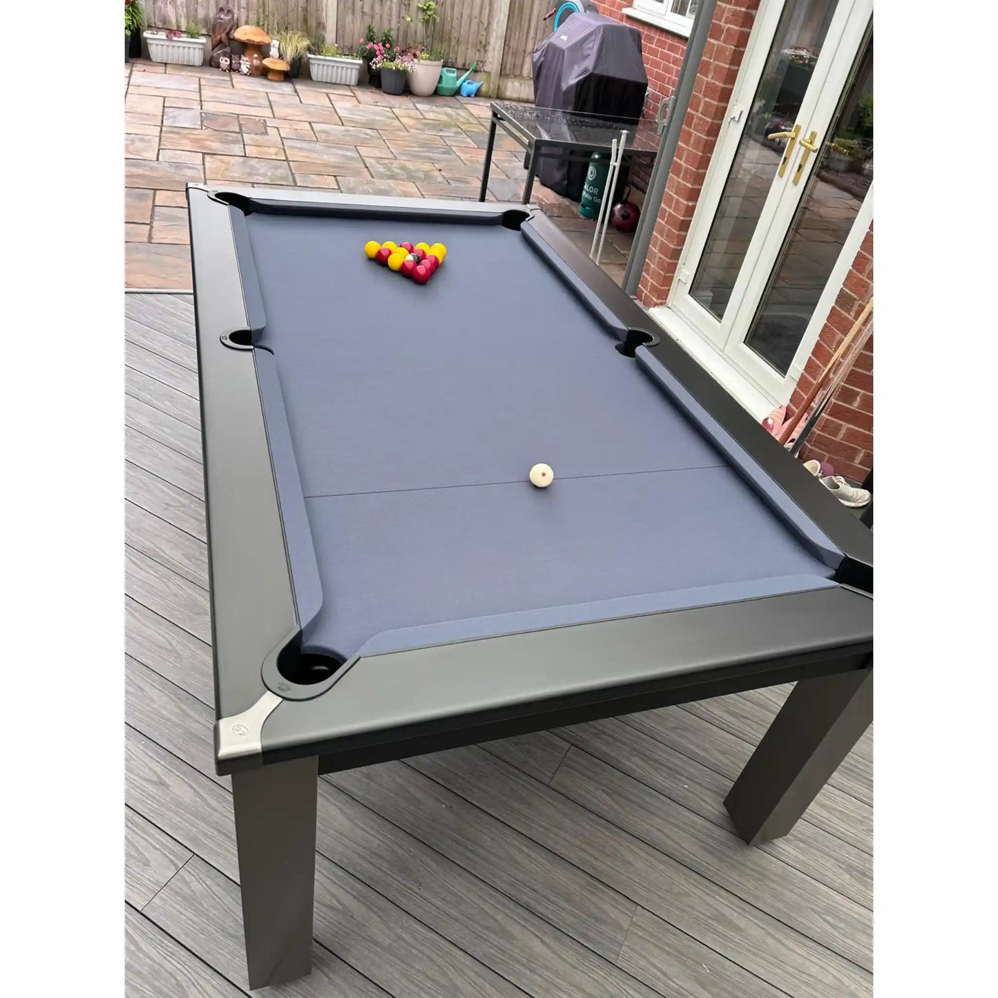 A grey pool table with slate blue felt is set up on a gray wooden deck in a backyard patio area next to a brick house.