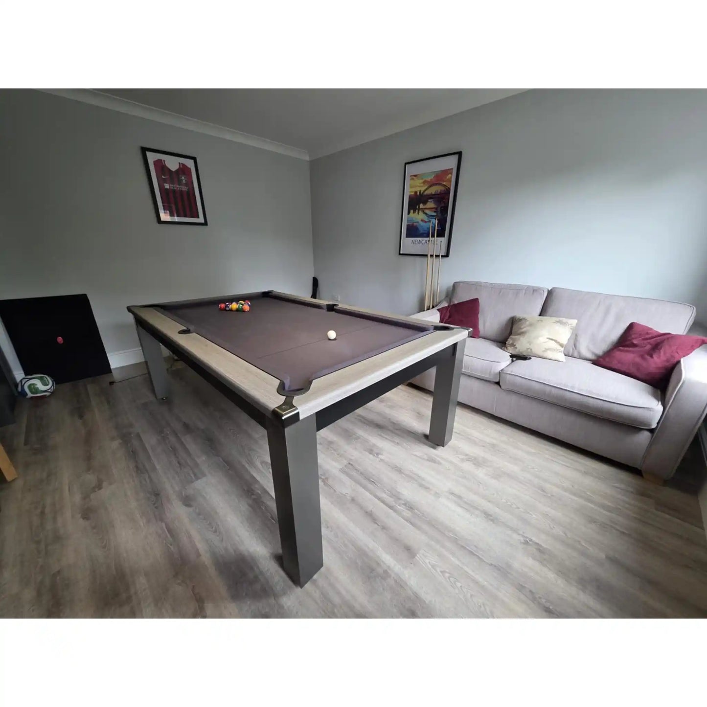 A rectangular pool table with a dark brown felt surface and light wood rails, positioned in a living room setting next to a light gray sofa with decorative pillows.