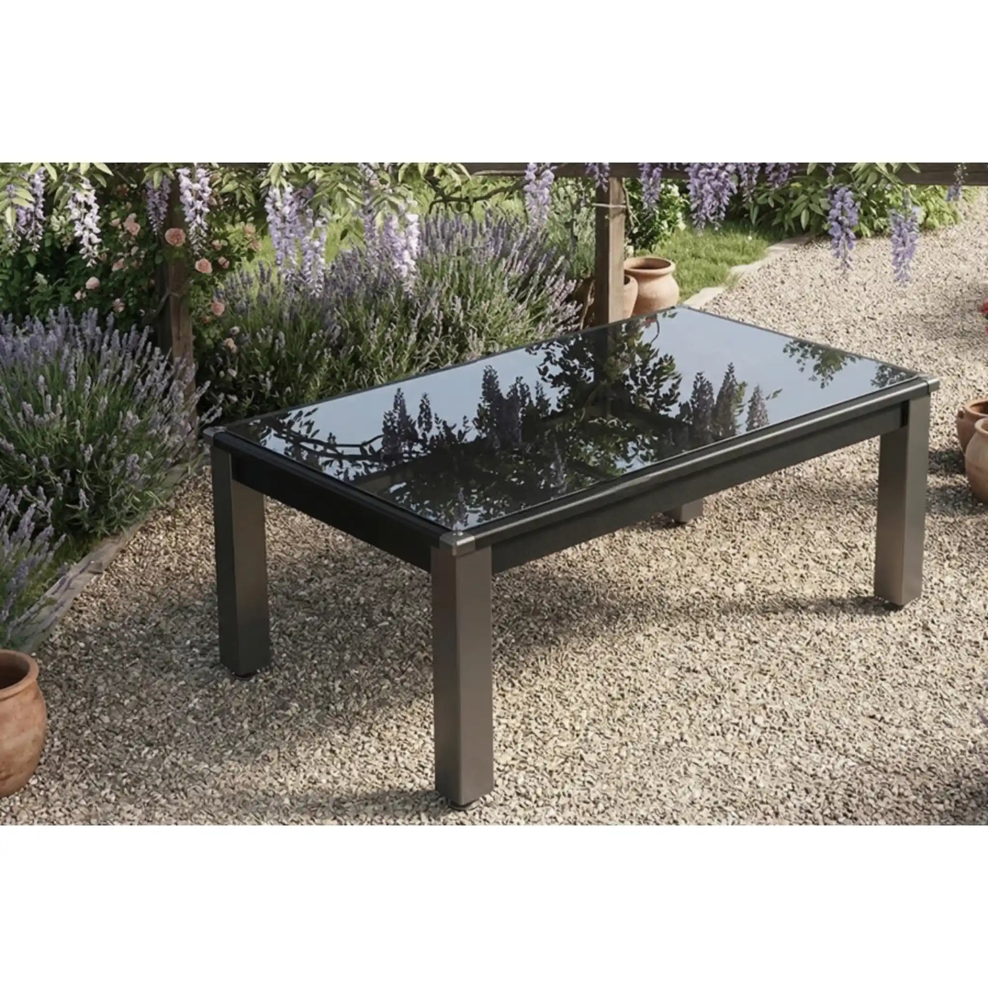 A rectangular outdoor pool dining table with a glossy black glass top and dark metal legs, set on a gravel patio in a garden with lavender and flowers in the background.