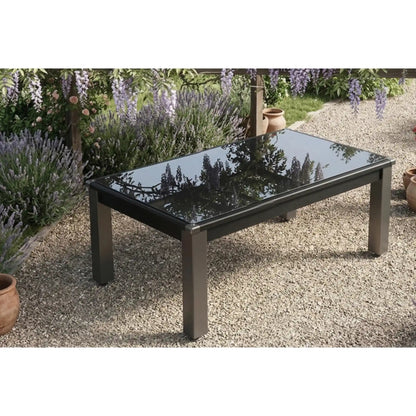 A rectangular outdoor pool dining table with a glossy black glass top and dark metal legs, set on a gravel patio in a garden with lavender and flowers in the background.
