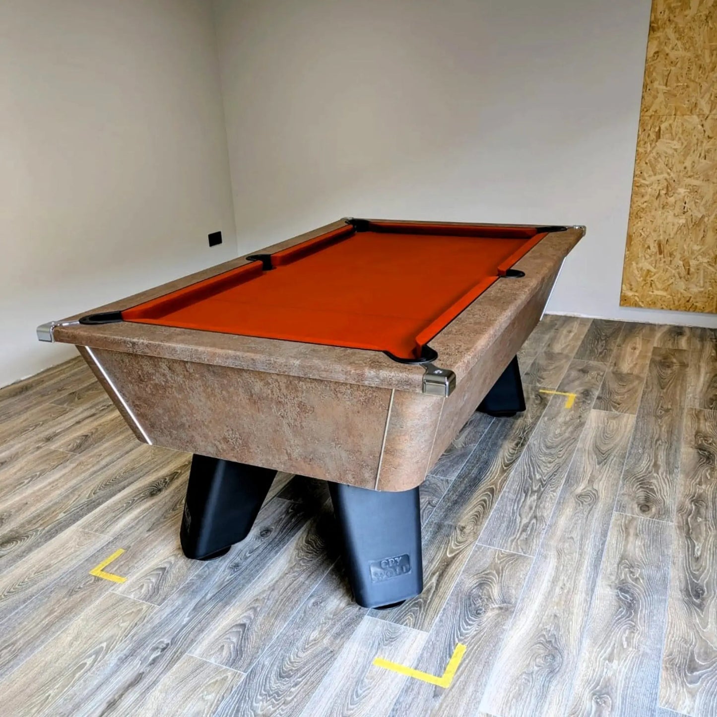 Pool table with orange felt on a wooden floor in a room with a plain wall.