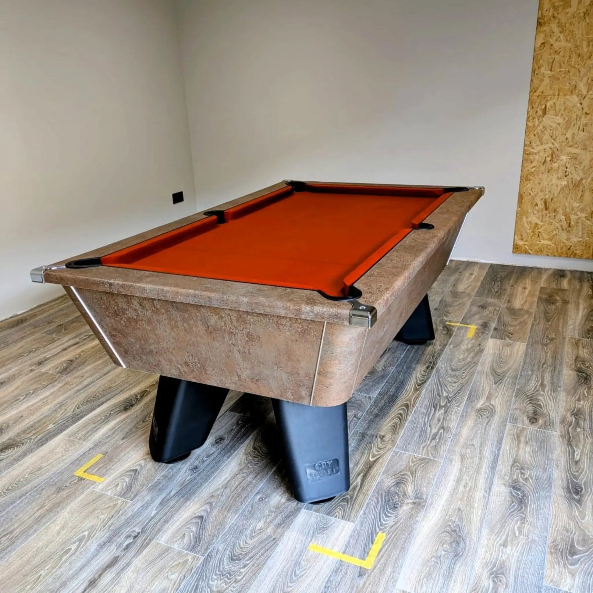 Pool table with orange felt on a wooden floor in a room with a plain wall.