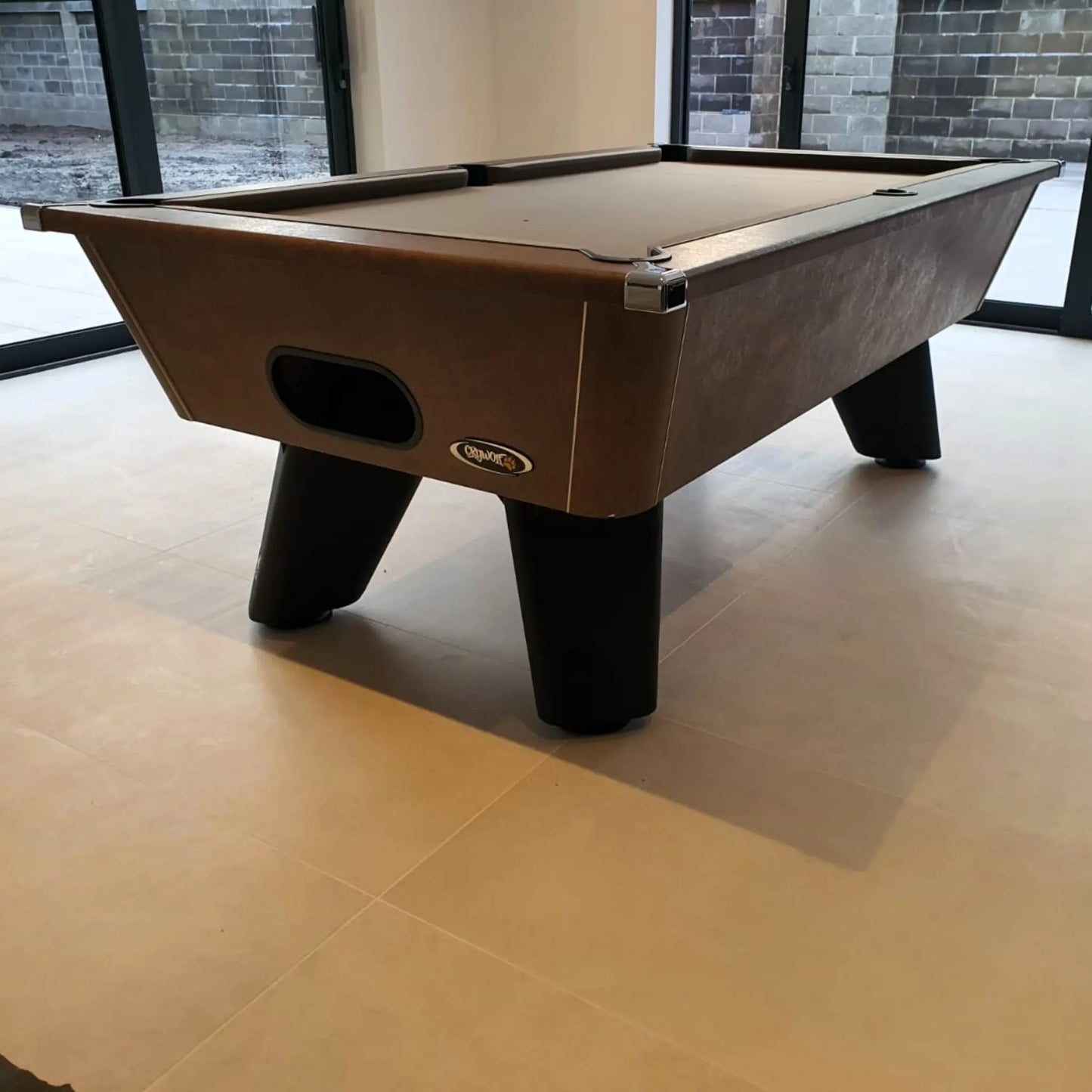 Pool table with brown felt surface and black legs in a room with large windows.