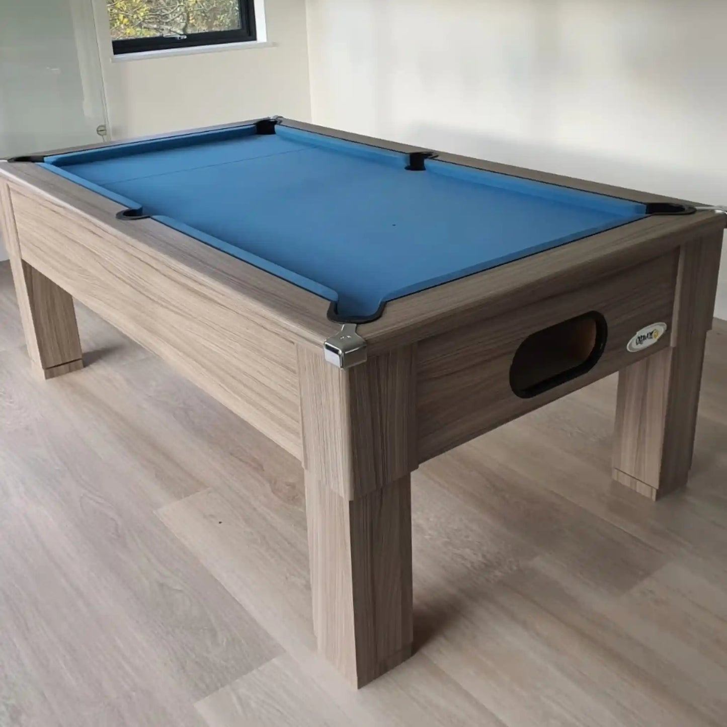 A wooden pool table with a bright blue felt playing surface, shown in a room with light wood flooring and off-white walls.
