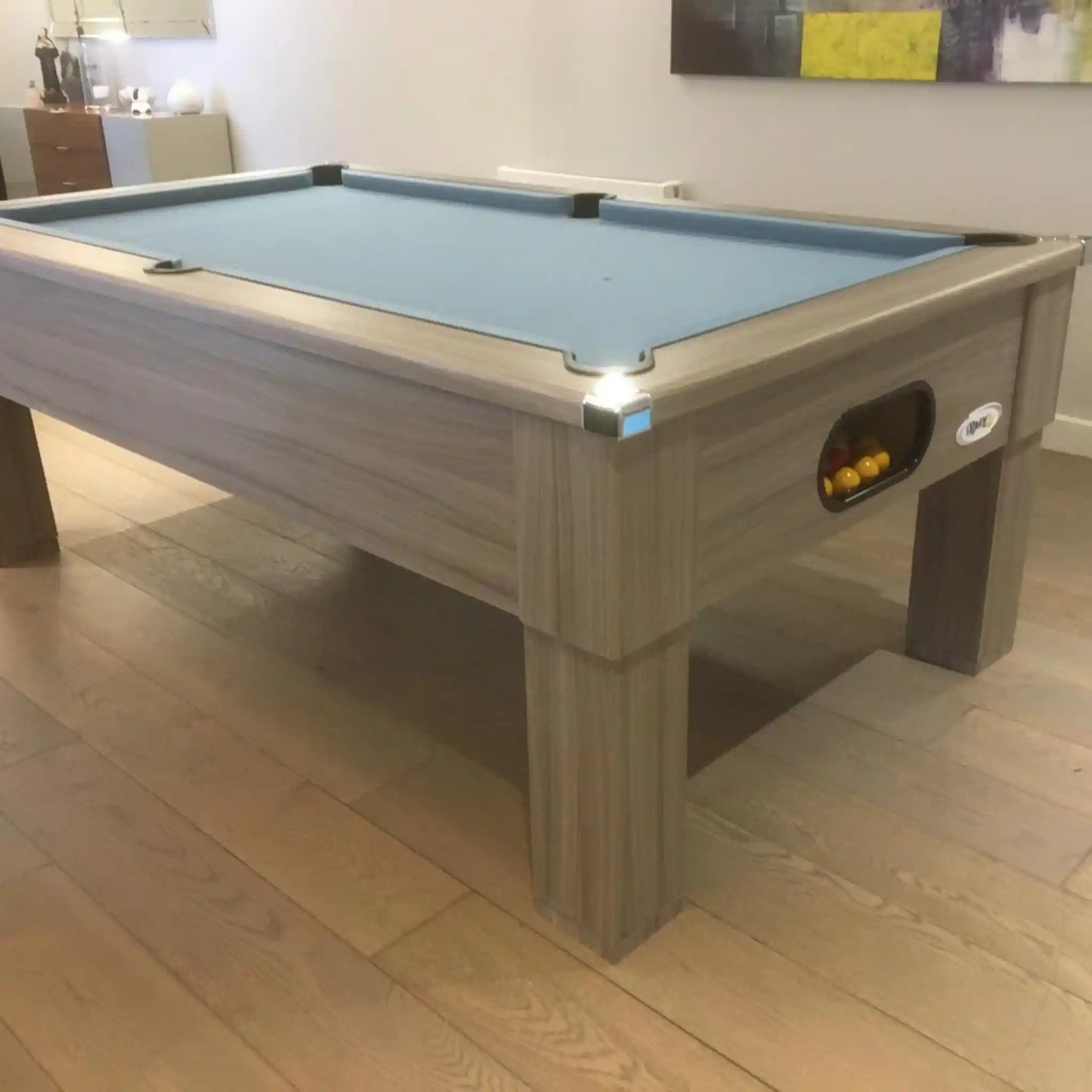 A wooden pool table with a light blue felt playing surface, shown in a real indoor room with wood flooring and wall decor visible.