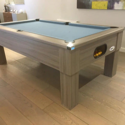 A wooden pool table with a light blue felt playing surface, shown in a real indoor room with wood flooring and wall decor visible.
