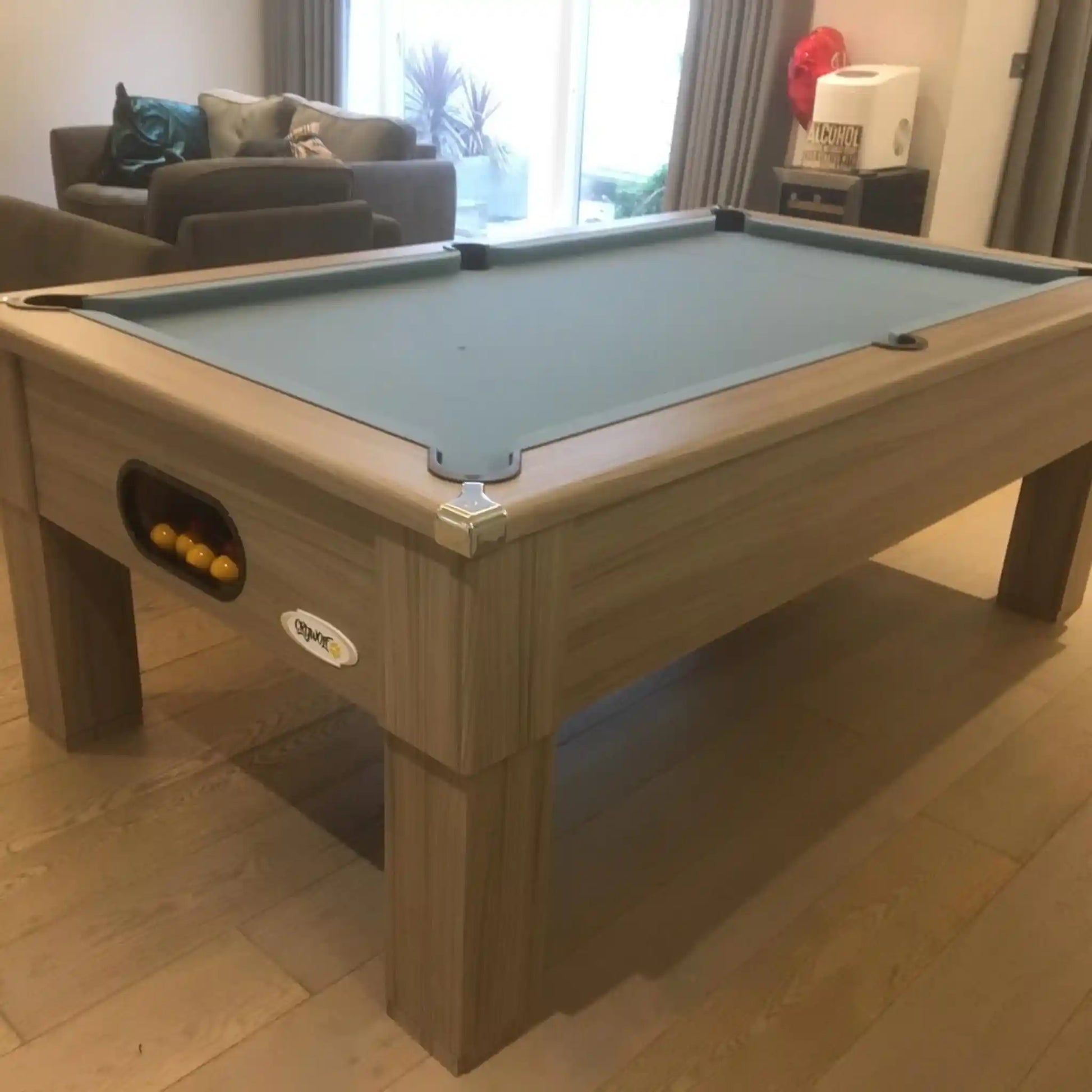 A wooden billiards table with a muted blue-green felt playing surface, shown in a living room setting with a sofa and window in the background.