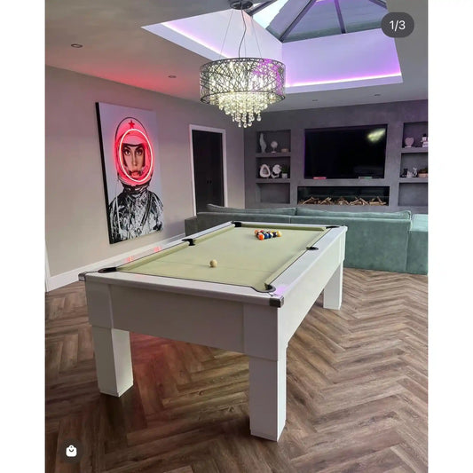 A white pool table with a light green felt surface in a modern living room setting with wood herringbone flooring, a crystal chandelier, and a wall art piece.