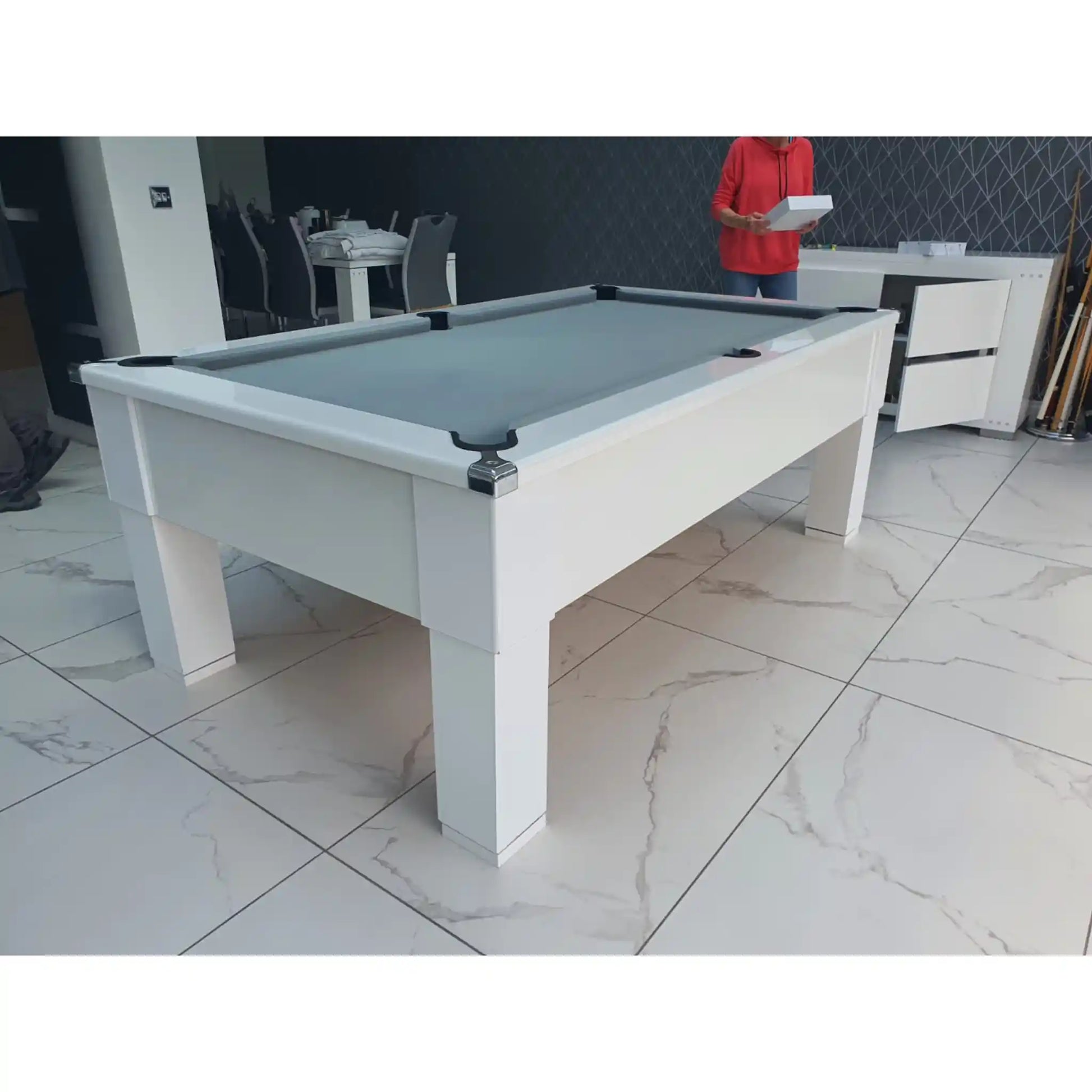 A modern white billiards table with a gray felt playing surface, shown in a showroom with a tiled floor and a person standing in the background.