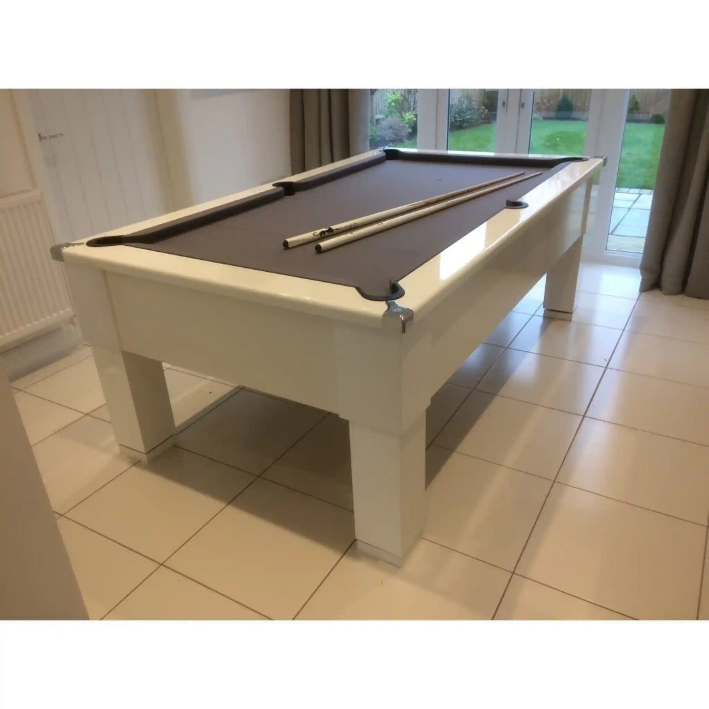 A white wooden pool table with a dark brown felt surface, shown in a tiled indoor room with large windows and a view of a green lawn outside.