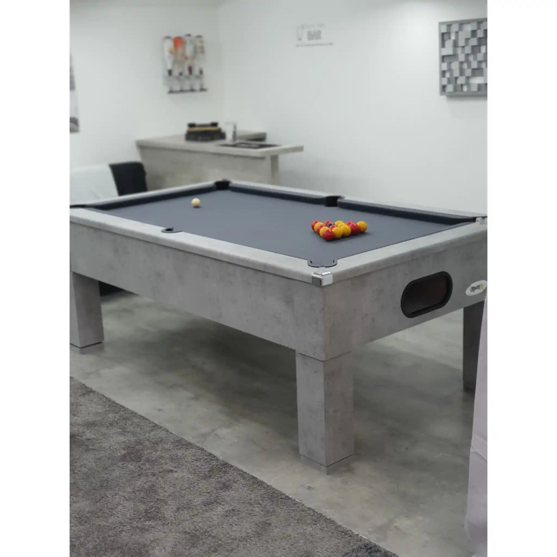 A modern pool table with a gray concrete-look frame and dark gray felt surface, shown in a minimalist indoor room with white walls and a gray rug.
