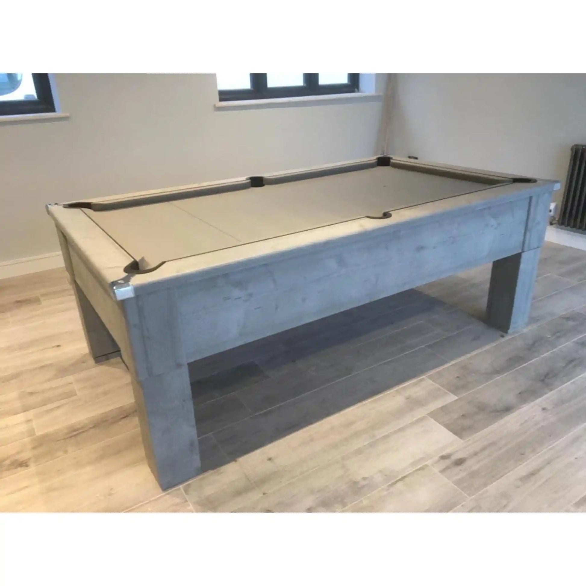A modern pool table with a light grey concrete-look frame and a beige felt playing surface, shown in a room with light wood floors and neutral walls.