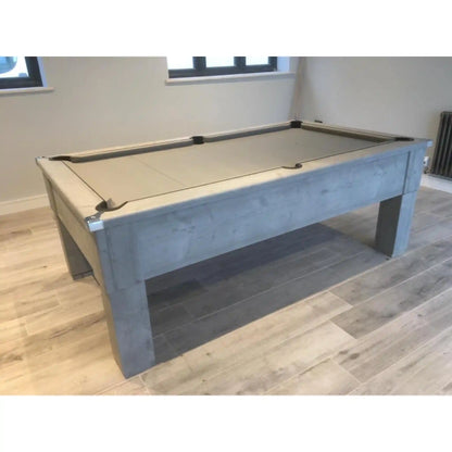 A modern pool table with a light grey concrete-look frame and a beige felt playing surface, shown in a room with light wood floors and neutral walls.