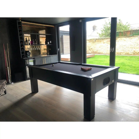 A black pool table with a dark felt surface is shown in a modern indoor room with large windows, a bar area, and a green backyard visible outside.