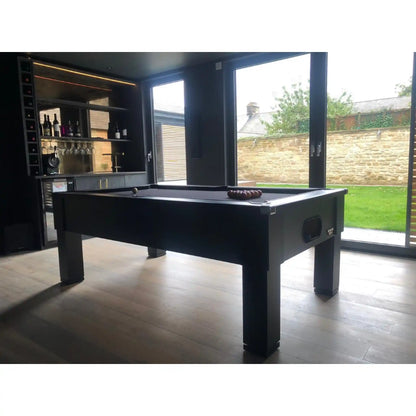A black pool table set in a modern, dark-themed indoor bar or lounge with large windows showing a backyard, a built-in bar shelf with bottles in the background, and a white border at the top and bottom of the image.