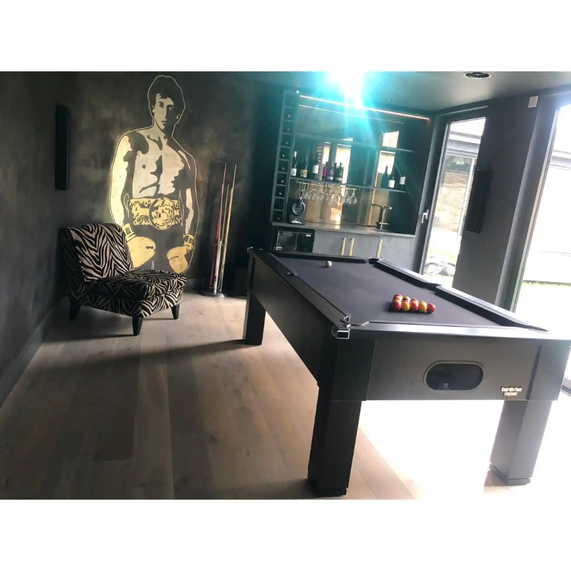 A dark wood pool table with a black felt surface and racked billiard balls, shown in a modern room with a zebra-print armchair, a large wall mural, and a bar cabinet in the background.