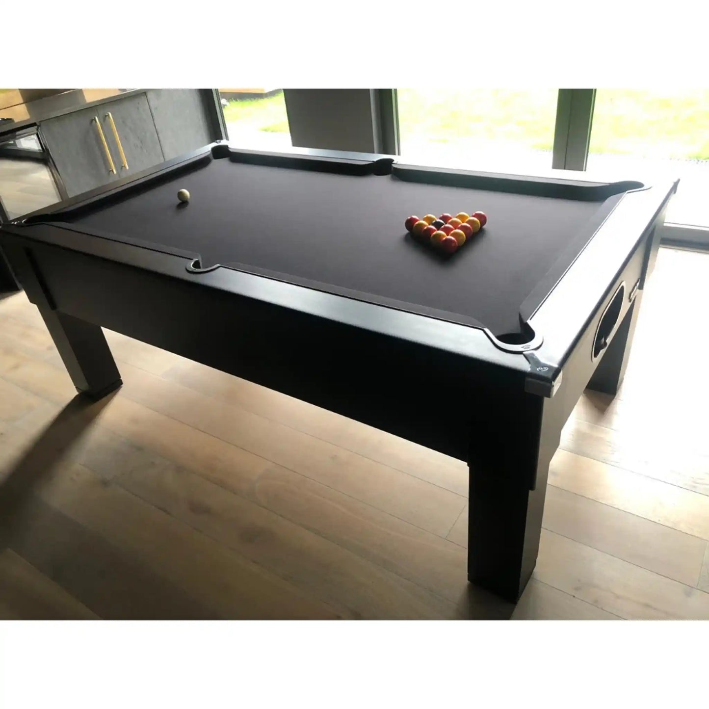 A black pool table with a black felt playing surface, shown in a room with light wood floors and windows in the background. A triangle of billiard balls and a cue ball are arranged on the table.