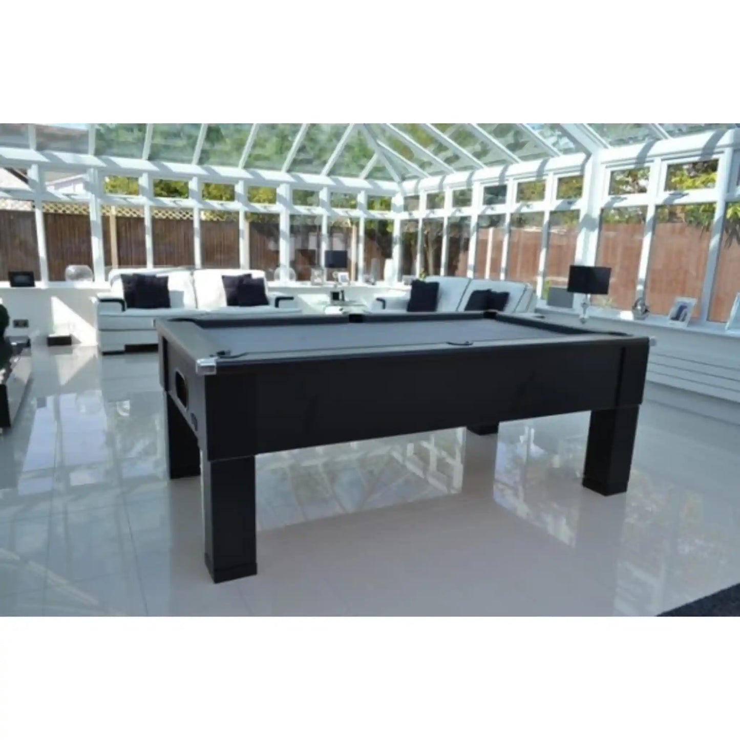 A black rectangular pool table positioned in a bright, modern conservatory with white-framed glass walls and white lounge furniture in the background.