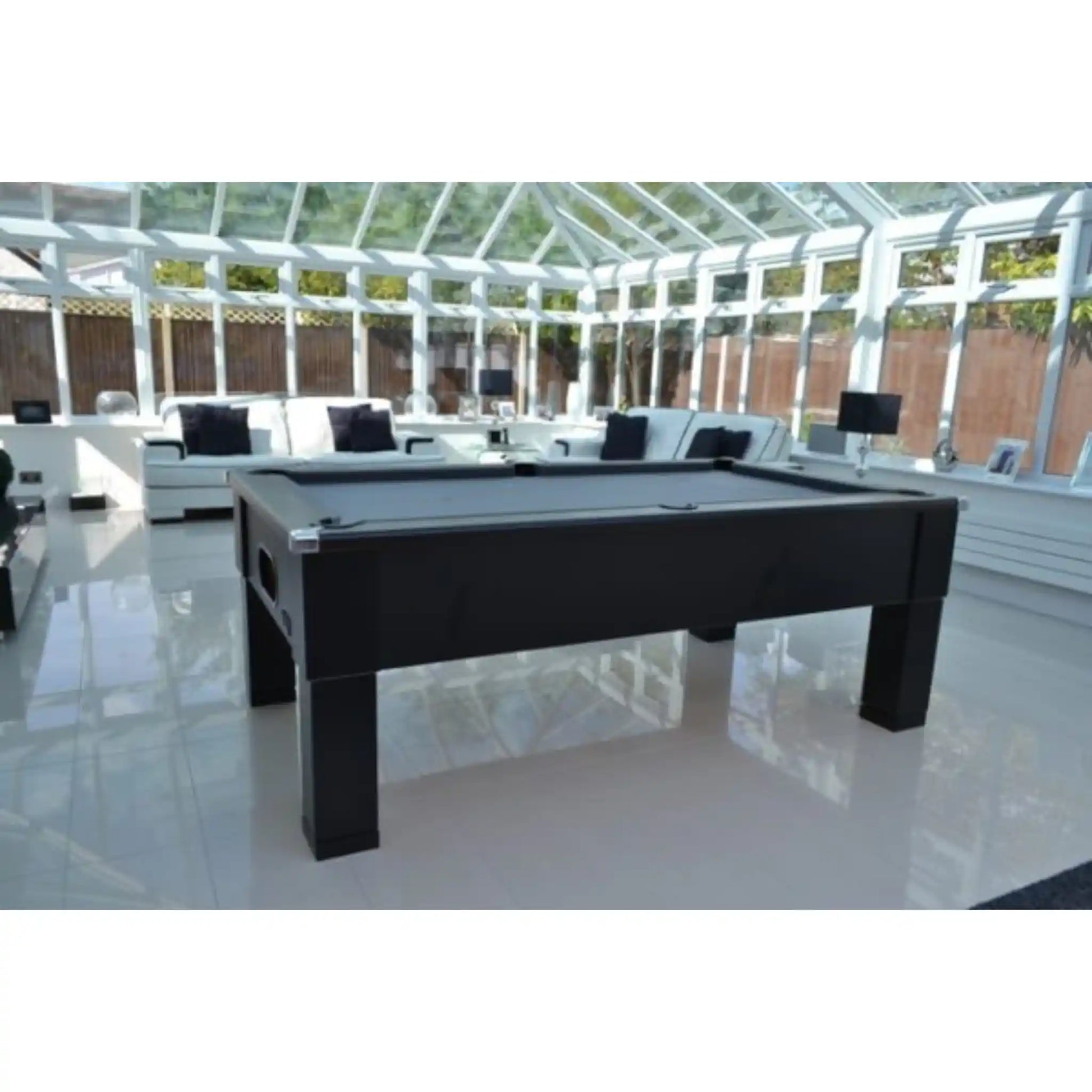 A black rectangular pool table positioned in a bright, modern conservatory with white-framed glass walls and white lounge furniture in the background.