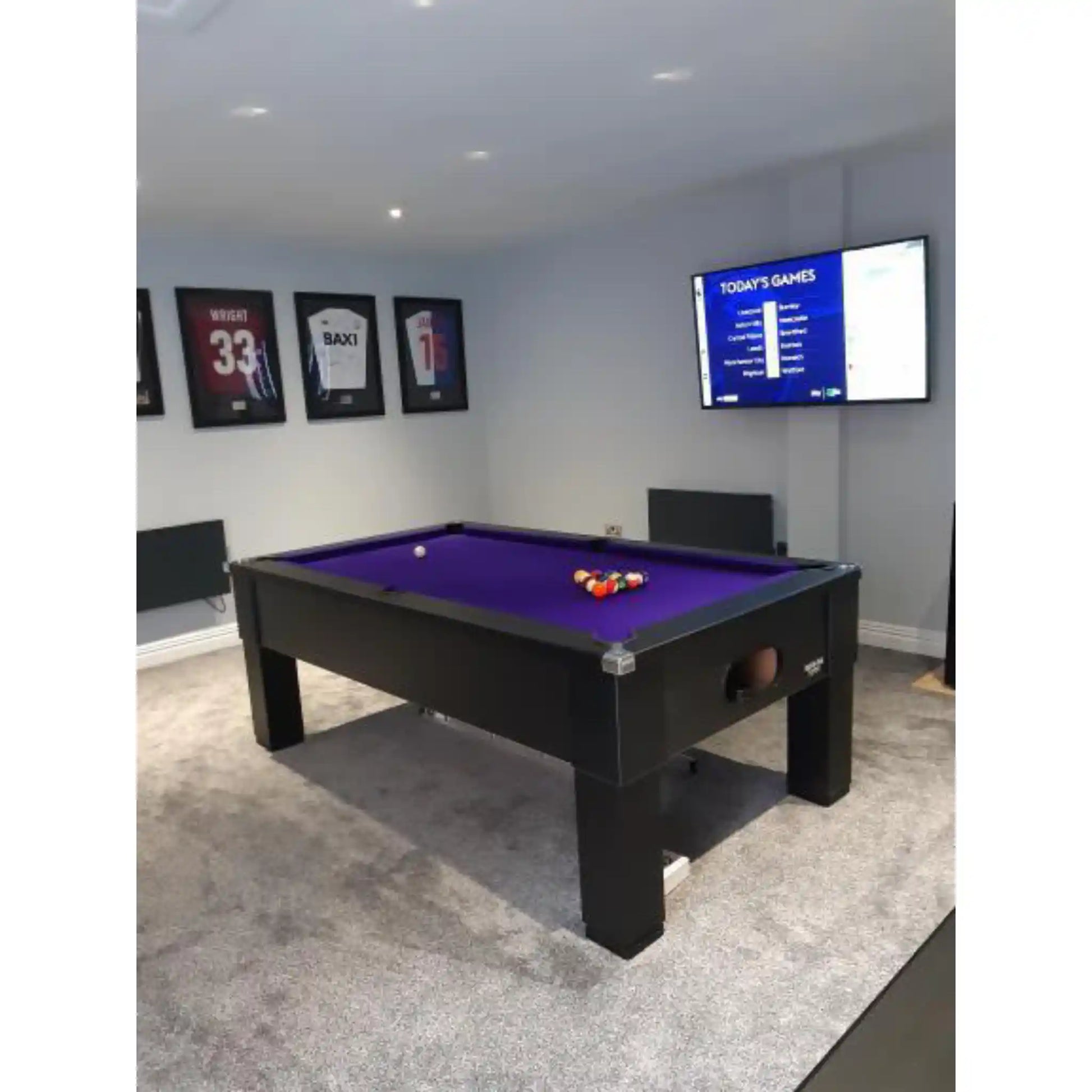A black pool table with a purple felt playing surface, set in a home entertainment room with gray carpet, framed sports jerseys on the wall, and a mounted TV screen.