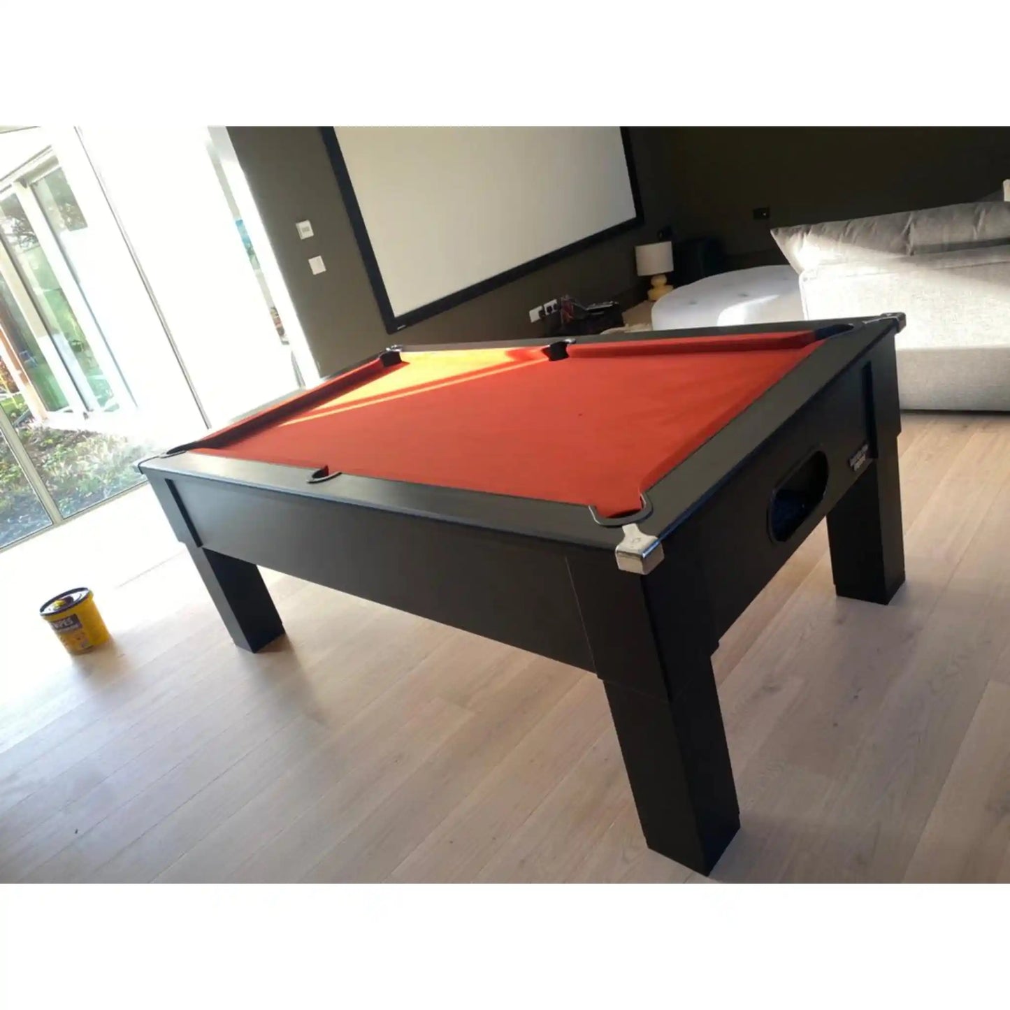 A modern pool table with a vibrant red felt surface and a dark charcoal gray wooden frame, shown in a living room setting with light wood floors.