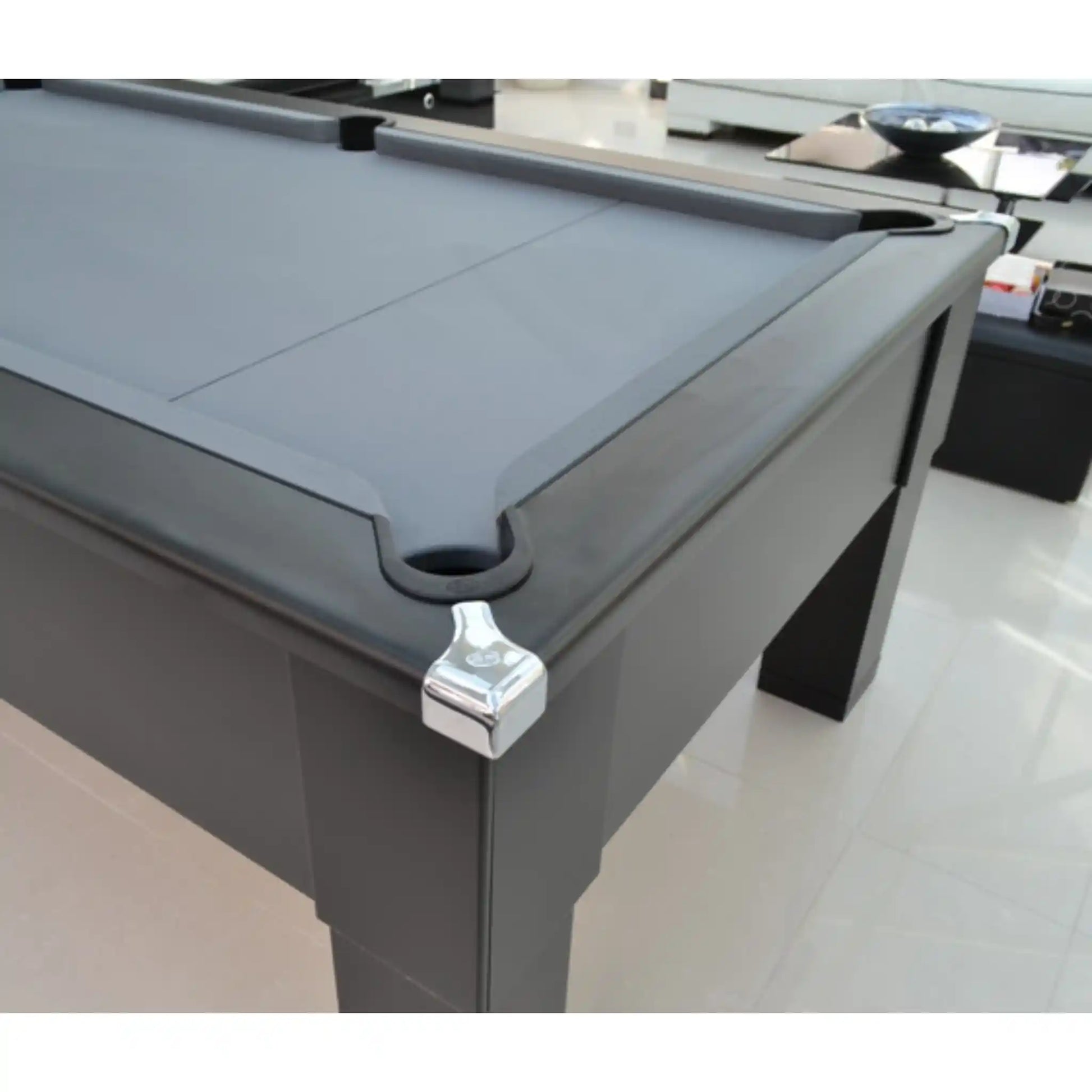 A close-up of a dark gray pool table corner with a white metal ball stopper and a black pocket, set in a bright modern room with light-colored tiled floors.