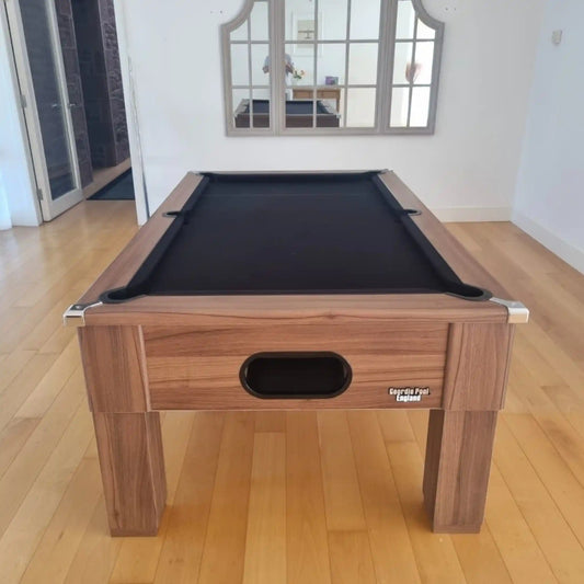 A wooden pool table with a black felt playing surface, photographed in a bright room with light wood floors and a mirror on the wall behind it.