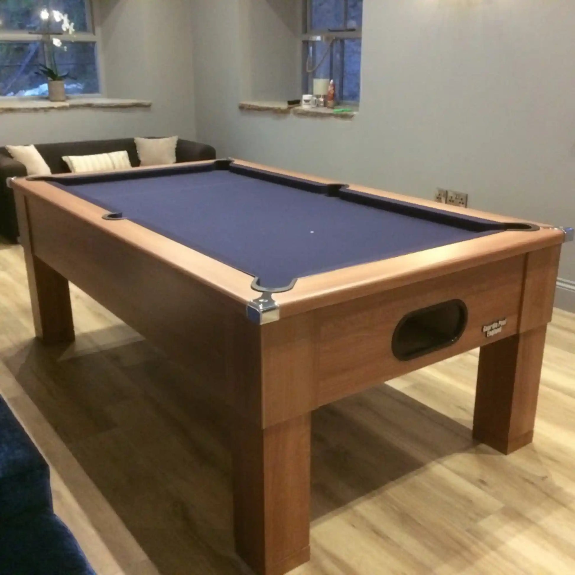 A wooden pool table with blue felt in a living room setting with a sofa, windows, and light wood flooring.