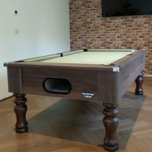 A dark wood pool table with a light green felt surface, shown in a room with a brick wall and wood parquet floor.