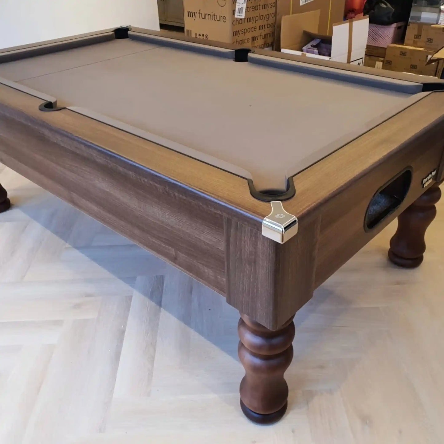 A wooden pool table with a grey felt playing surface, with cardboard boxes in the background.