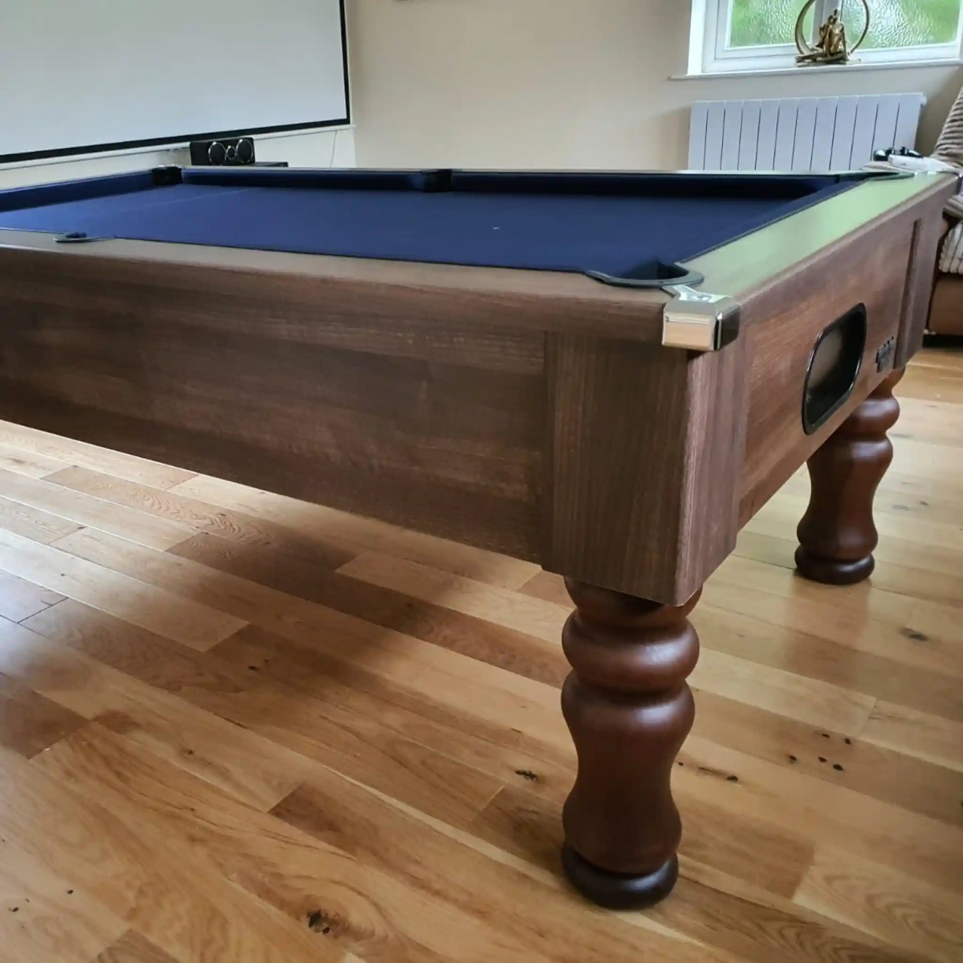 A wooden pool table with a dark blue felt playing surface, shown in a home environment with light wood floors and a window in the background.