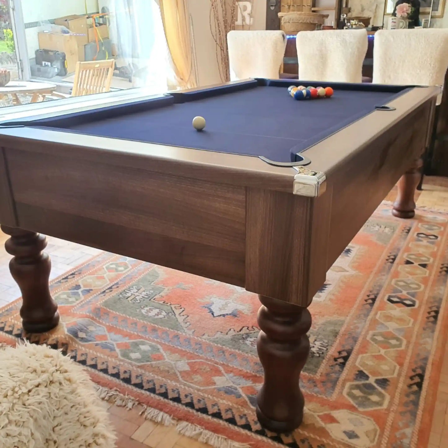 A wooden pool table with a dark blue felt surface, shown in a furnished room with chairs, a rug, and a billiard ball set on top.