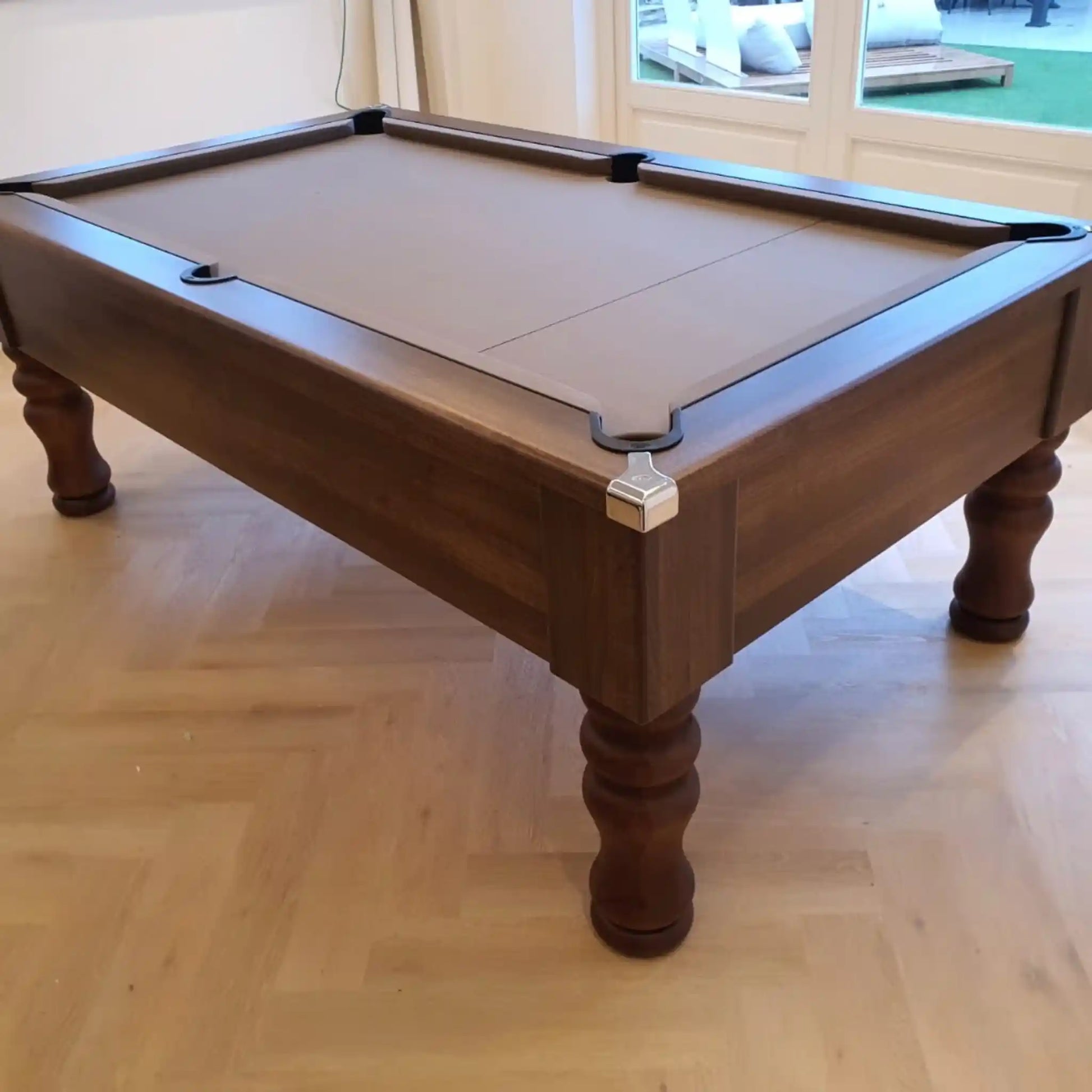 A dark brown wooden pool table with a matching felt-covered playing surface, shown in an indoor room with light wood flooring and a window in the background.