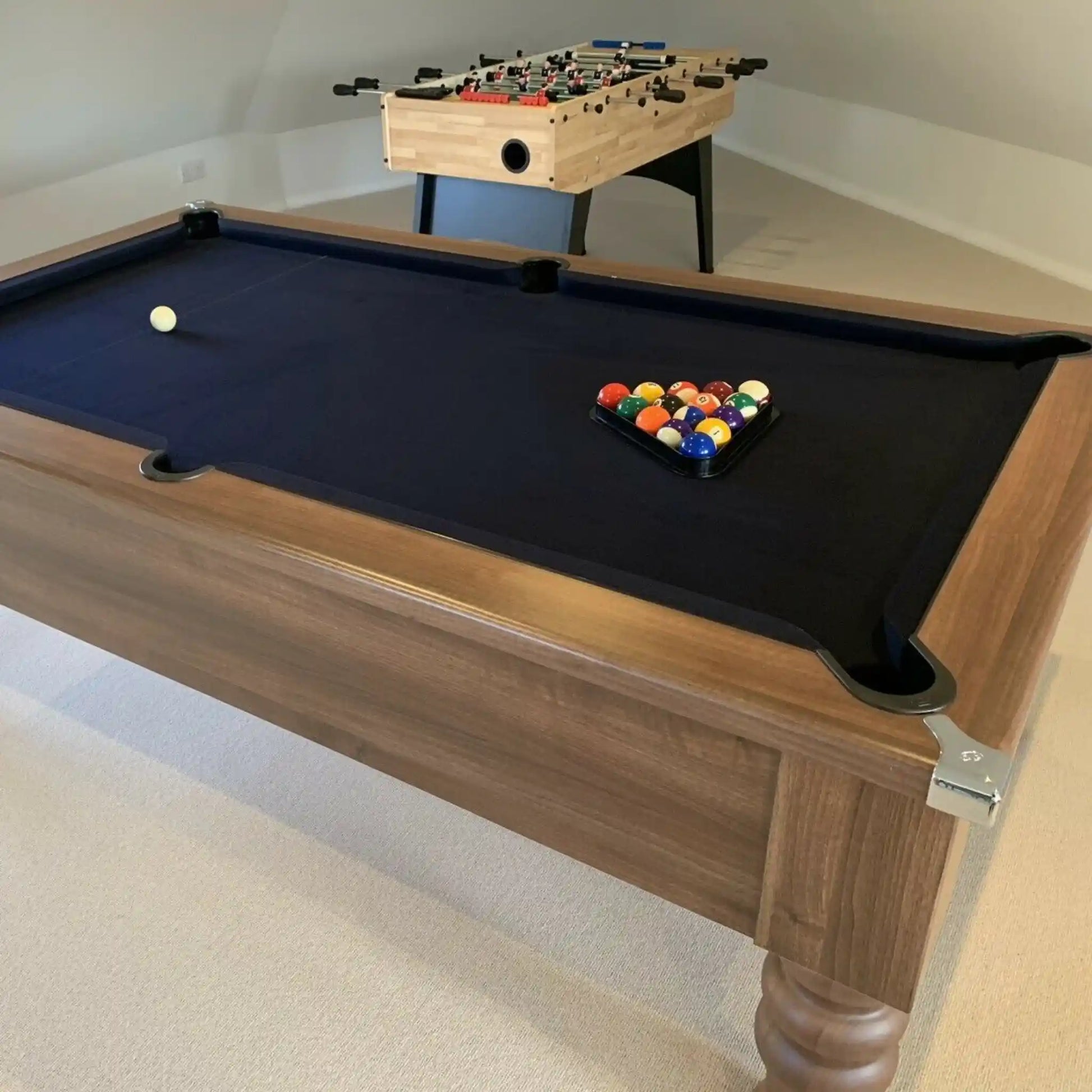 A wooden pool table with a black felt surface, shown in a home setting with a foosball table in the background.