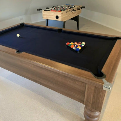 A wooden pool table with a black felt surface, shown in a home setting with a foosball table in the background.