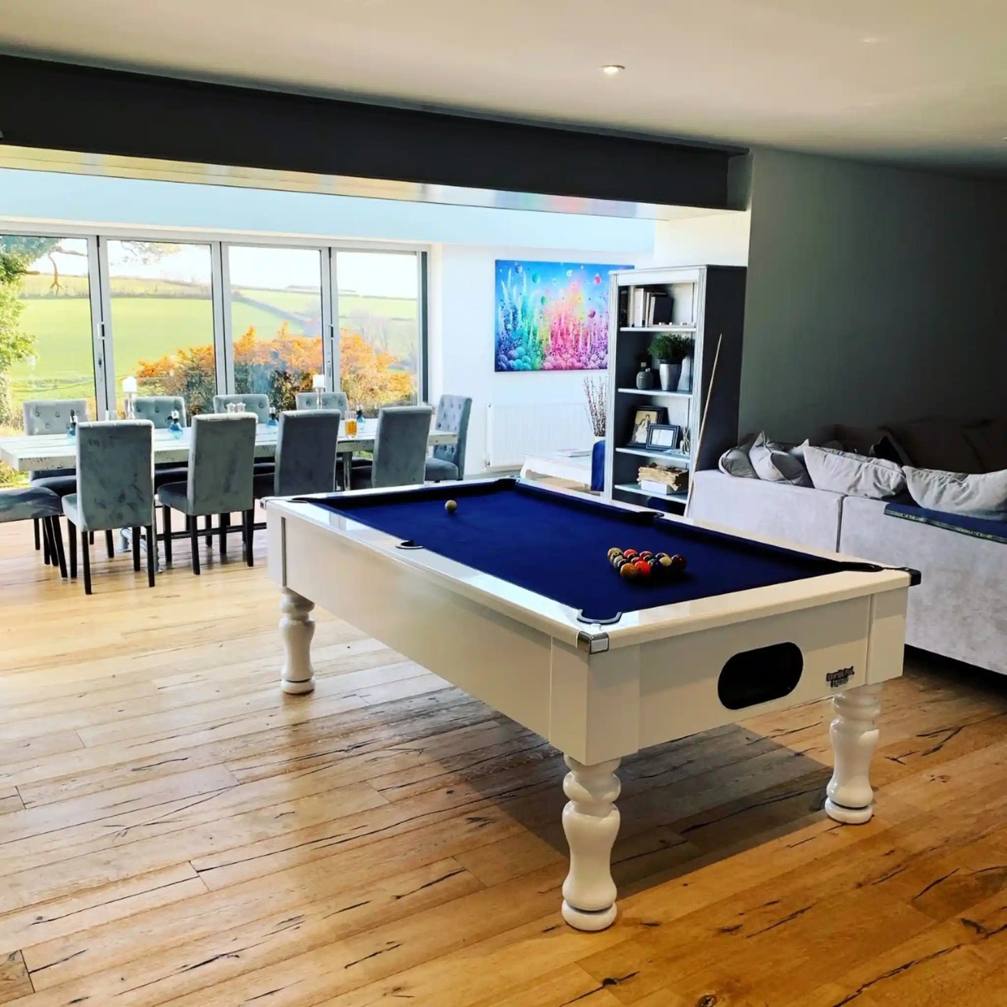 A white pool table with a deep blue felt surface is shown in a modern living room with hardwood floors, a dining area, and a sofa in the background.