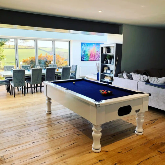 A white pool table with a deep blue felt surface is shown in a modern living room with hardwood floors, a dining area, and a sofa in the background.