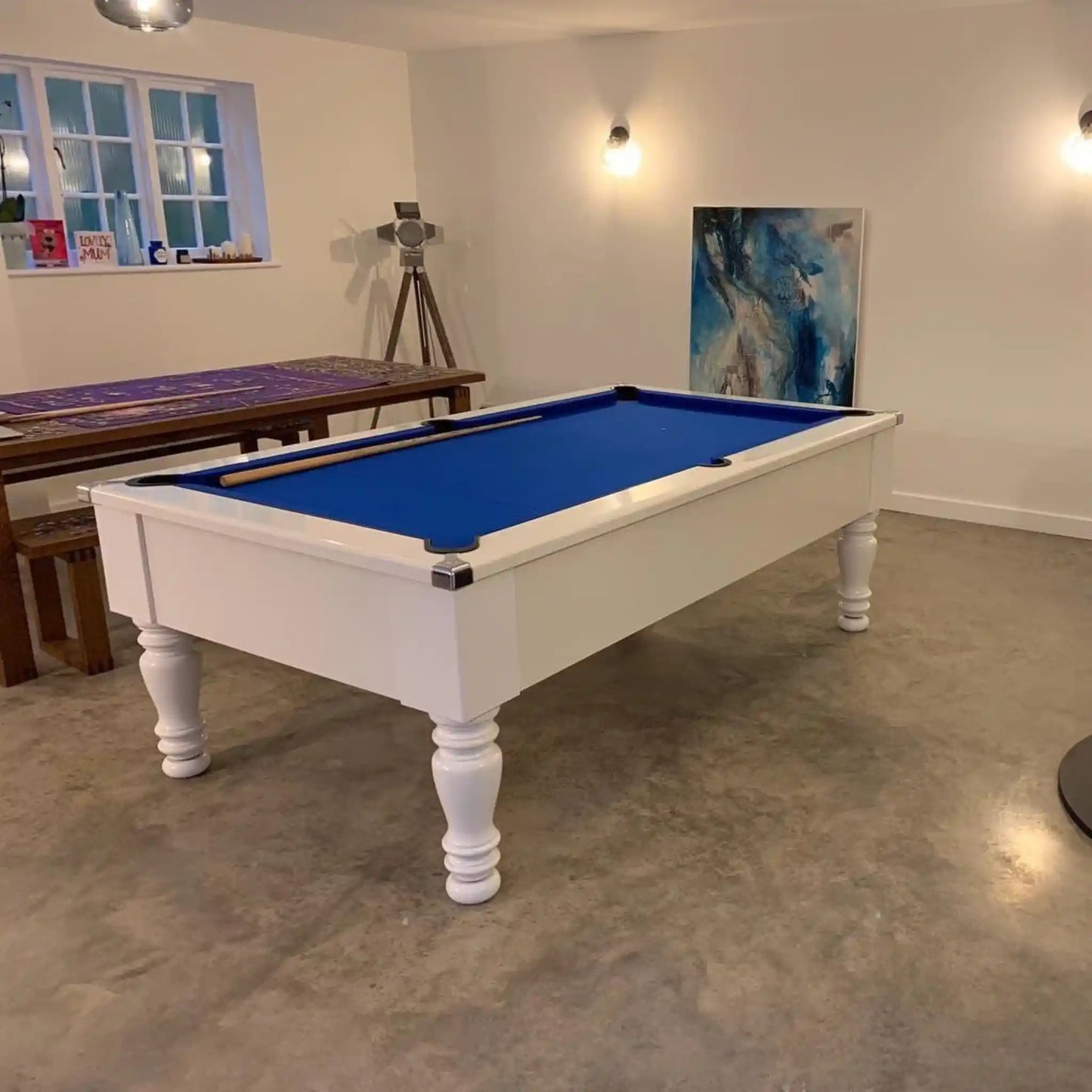 A white wooden pool table with a vibrant blue felt playing surface, positioned in a modern room with concrete floors and white walls.