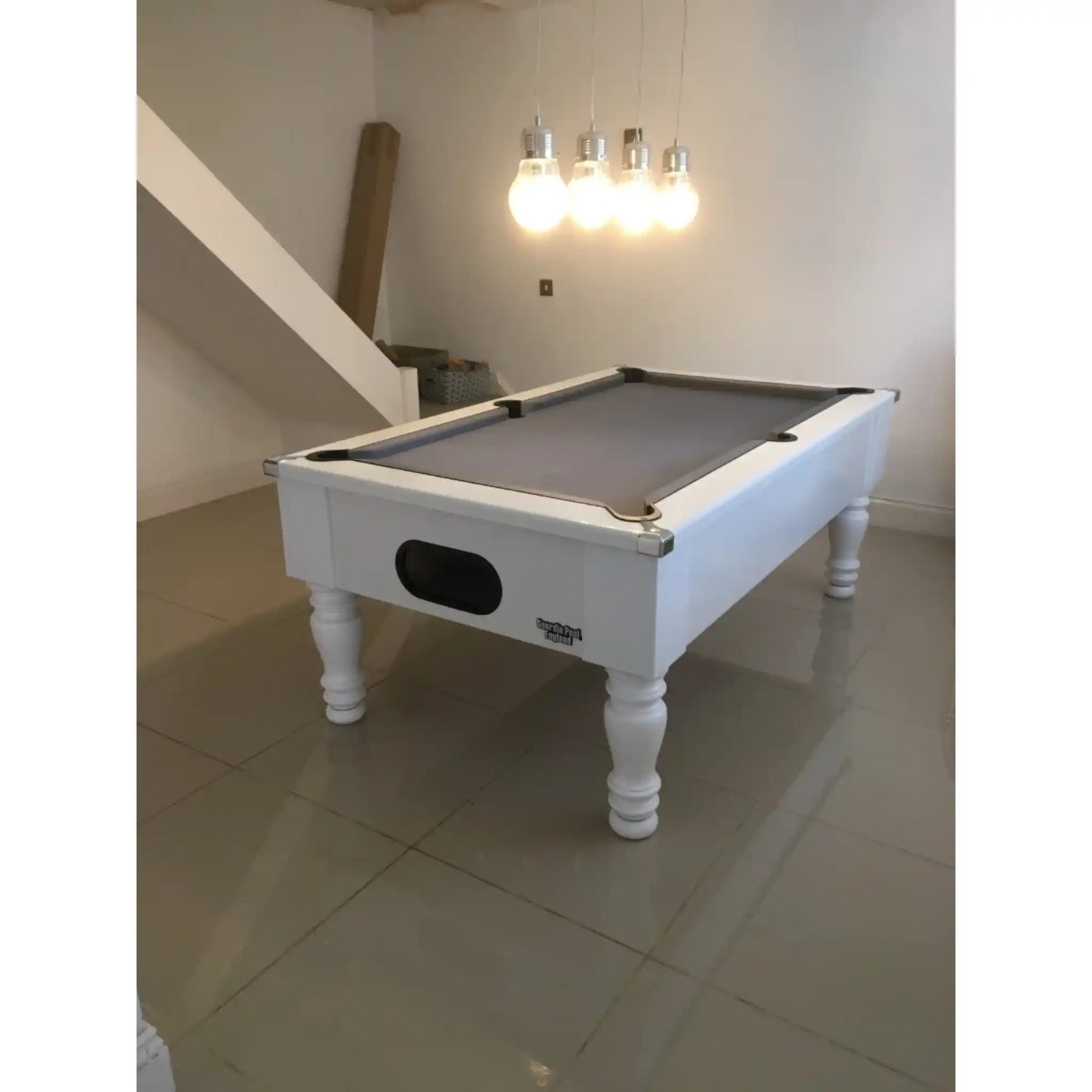 A white wooden pool table with a grey felt playing surface, shown in a room with tiled floors, a staircase, and hanging light bulbs.