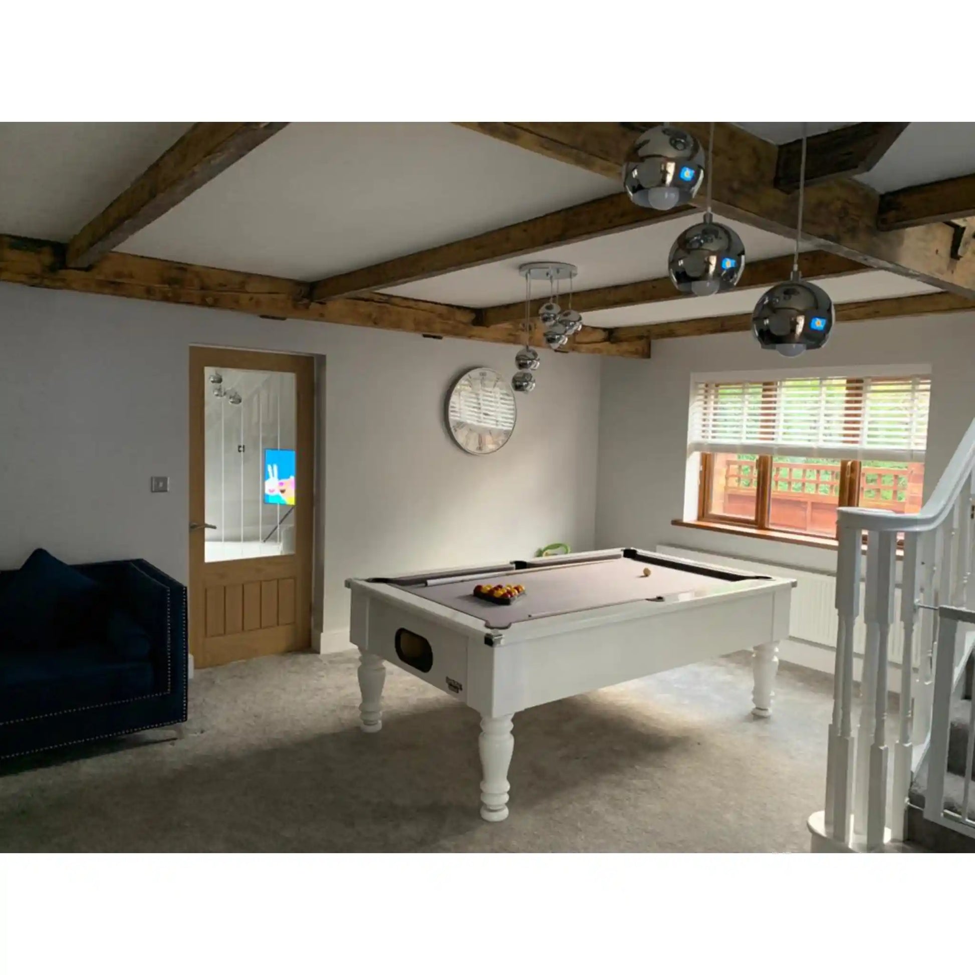 A white wooden pool table with a purple felt surface in a bright, modern room with exposed wooden ceiling beams, three silver globe pendant lights, a blue armchair, and a staircase on the right.
