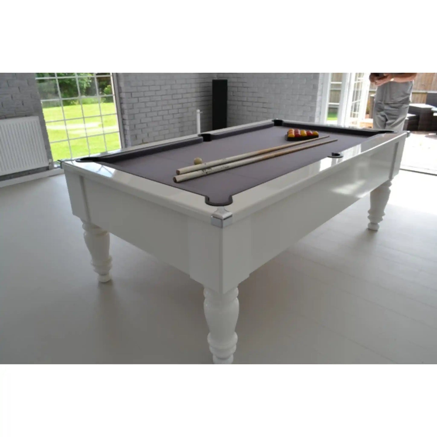 A white wooden pool table with a dark grey felt surface, shown in a bright modern room. A set of billiards, including cues and balls, is arranged on the table, and a person is partially visible in the background near the windows.