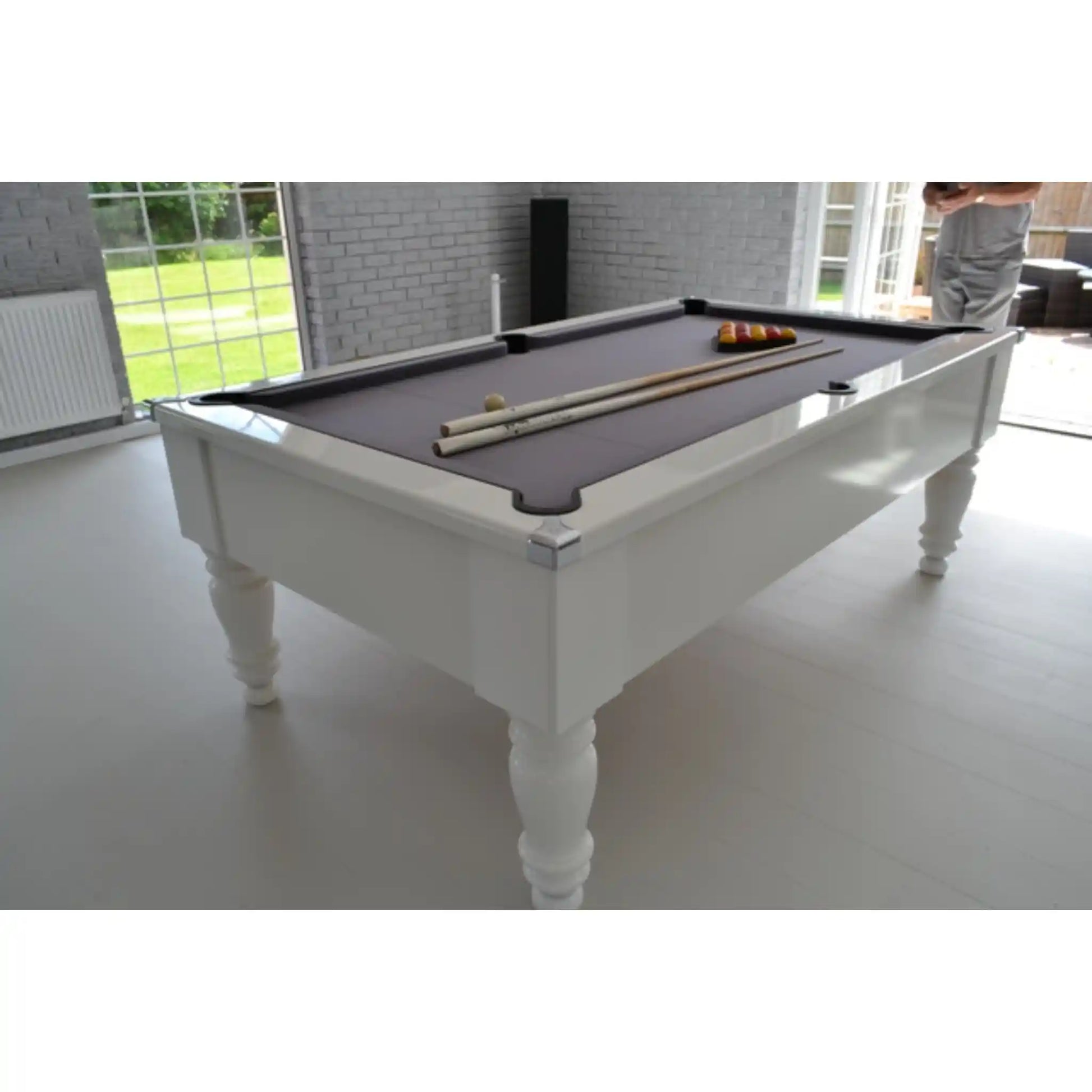 A white wooden pool table with a dark grey felt surface, shown in a bright modern room. A set of billiards, including cues and balls, is arranged on the table, and a person is partially visible in the background near the windows.