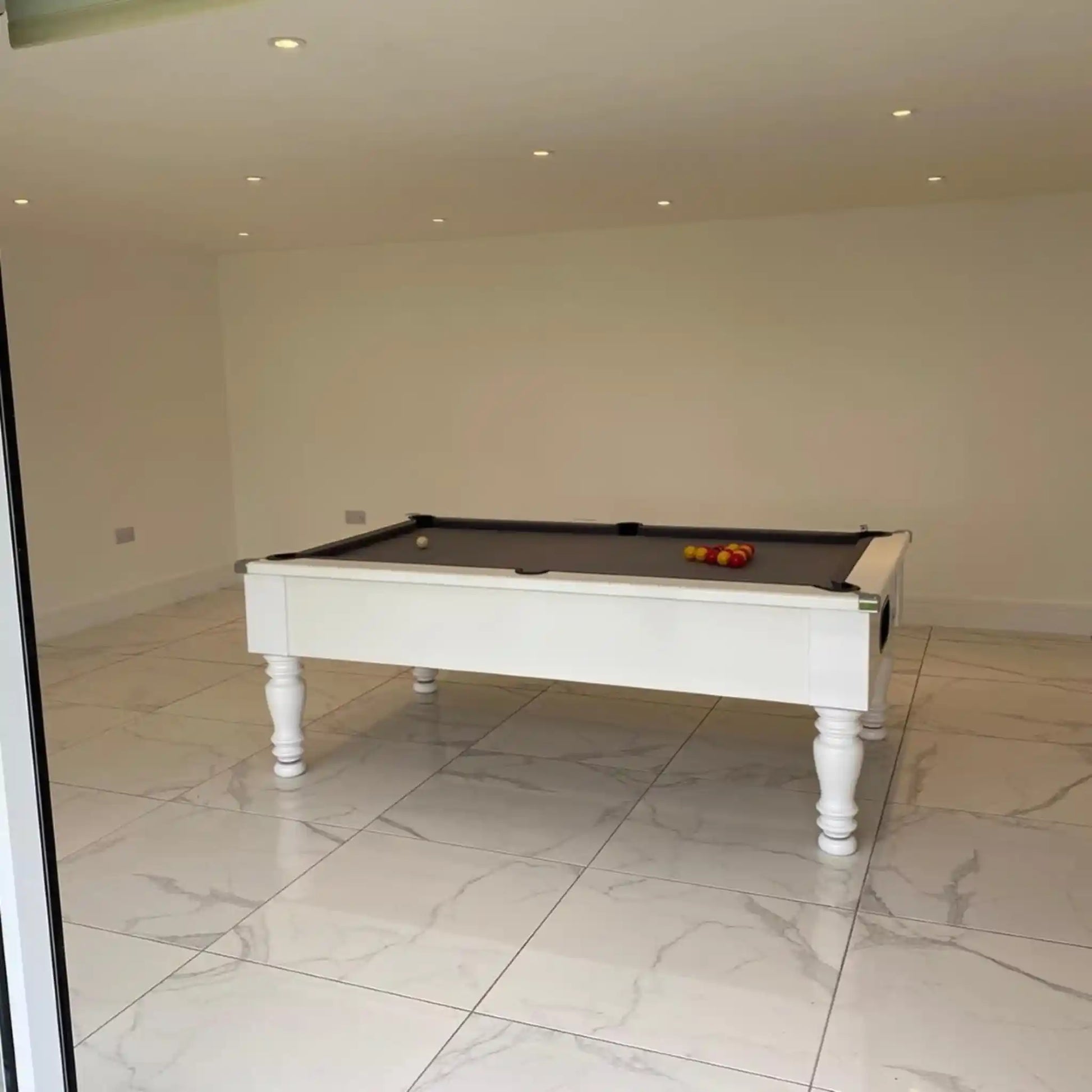 A white pool table with a dark felt surface and a few colored balls on top, set in a spacious room with light beige walls and large white marble-look floor tiles.