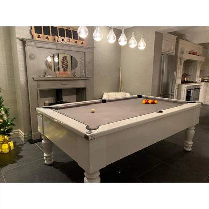 A white wooden billiards table with a gray felt playing surface, shown in a residential living room setting with a fireplace, pendant lights, and kitchen cabinets in the background.
