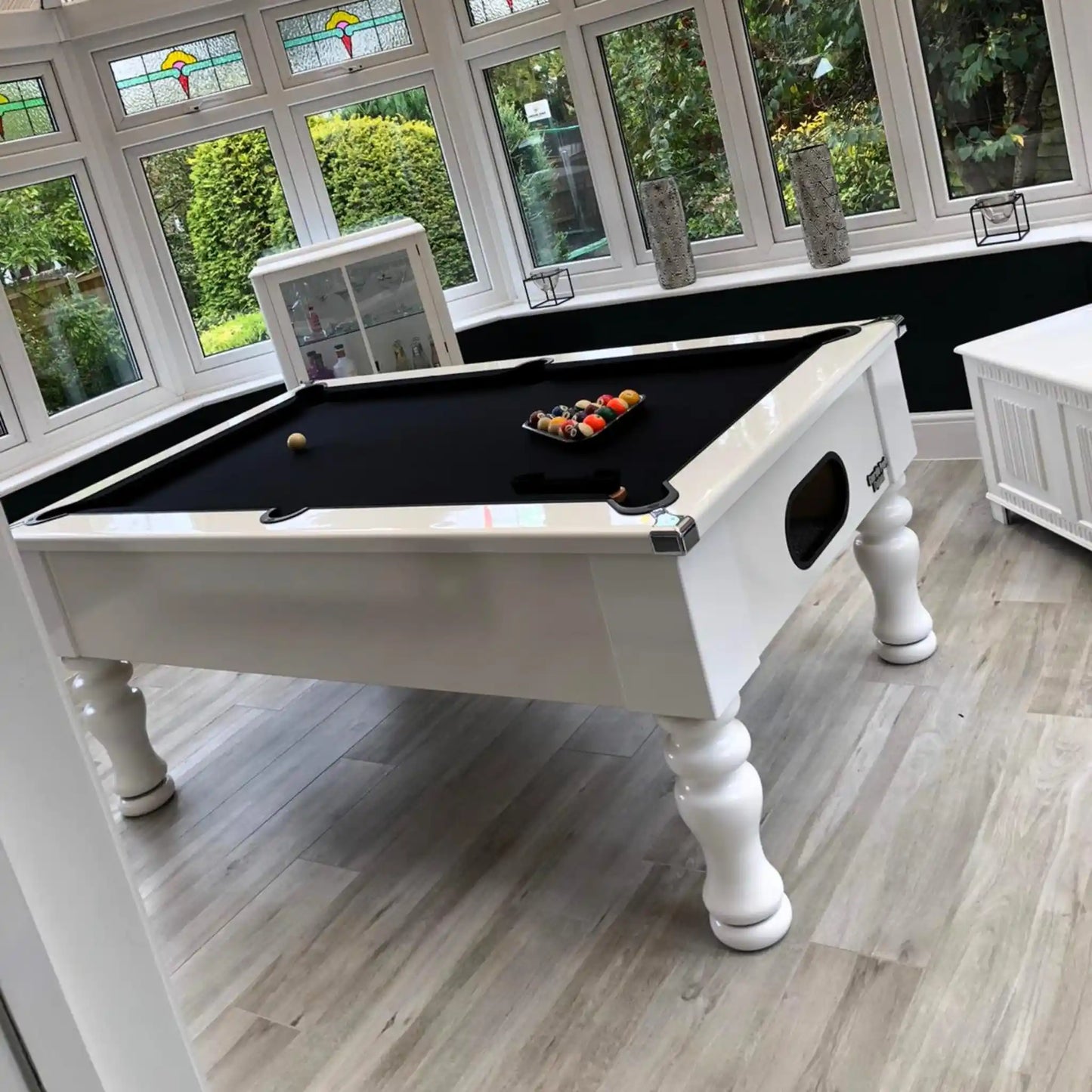 A white wooden pool table with a black felt playing surface, shown in a bright sunroom with large windows and light wood flooring.