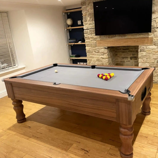 A wooden pool table with a grey felt surface and a triangle rack of yellow and red billiard balls, photographed in a furnished room with wood floors, a stone fireplace, shelving, and a mounted TV.